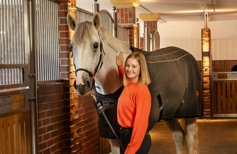 A horse stands in the stable next to its rider, wearing the St. Moritz heated blanket.