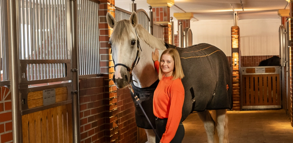 A horse stands in the stable next to its rider, wearing the St. Moritz heated blanket.