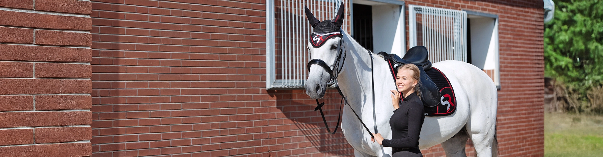 Eine Reiterin steht neben einem Pferd mit schwarz roter Schabracke und passender Fliegenhaube mit SR Logo vor einem Stall