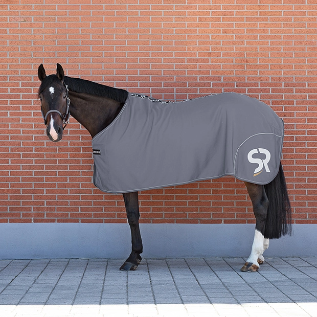 Ein Pferd steht vor einer roten Ziegelwand und trägt eine graue Abschwitzdecke Boston von Sunride