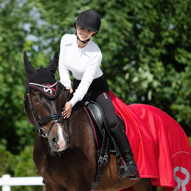 Eine Reiterin sitzt auf einem Pferd und streichelt dessen Hals Das Pferd trägt eine rote Abschwitzdecke Boston und passende Fliegenohren von Sunride