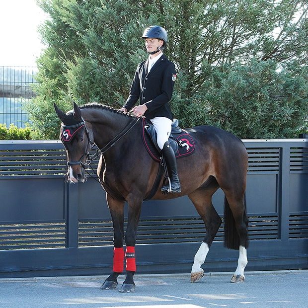 Ein Reiter sitzt auf seinem Pferd, das mit Trense, Schabracke und roten Bandagen von Sunride ausgestattet ist
