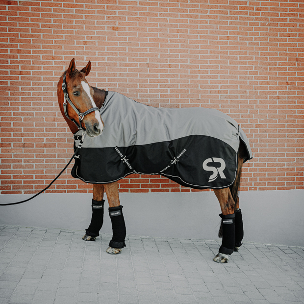 horse with black leather halter dallas, winter rug helsinki 250 grams grey and stable bandages black leather halter dallas and black stable bandages with pads and grey helsinki winter rug with 250 grams filling and reflecting elements and red leather cord halter by sunride on a horse in front of a stable