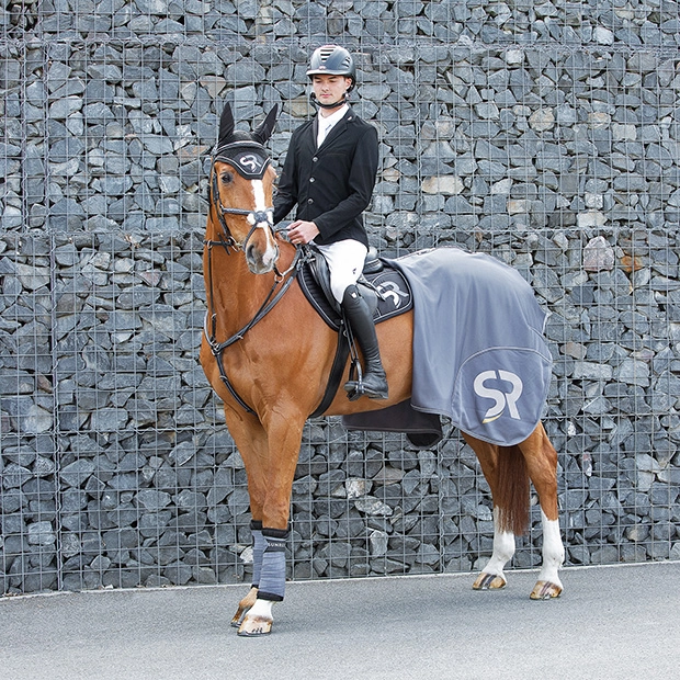 1 Ein Reiter sitzt auf einem Pferd vor einer Steinmauer Das Pferd trägt eine graue Abschwitzdecke Boston von Sunride sowie passendes Zubehör